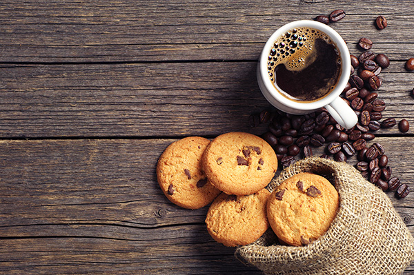 white mug of coffee on a dark wood surface. Loose coffee beans and a bag of chocolate chip cookies surround the mug.