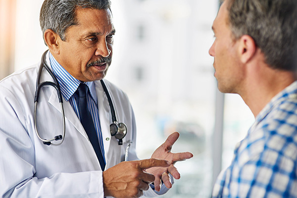 a doctor wearing a lab coat and stethoscope speaks to a patient in a blue shirt.